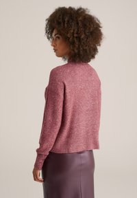 Femeie cu păr creț, purtând un pulover roz cu stropi și o fustă din piele violet, cu talie înaltă, stând de profil pe un fundal simplu.