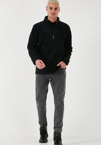 Pullover in fleece nero con collo a mezza zip, dotato di maniche lunghe, una texture liscia e una cucitura orizzontale in contrasto. Abbinato a jeans grigi e scarpe nere.