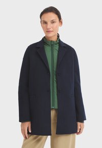 Abrigo de lana azul marino con solapas estructuradas y dos botones, superpuesto a una chaqueta verde con cremallera. Pantalones marrón claro. Textura suave, corte ajustado.