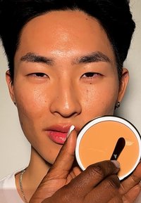 Krämig foundationkompakt med en slät, beige yta, hålls av en hand med mörk hy, nära en models ansikte som visar hudens textur.