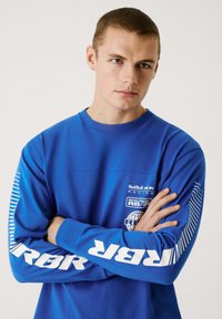 Blå sweatshirt lavet af bomuld, med hvide grafiske logoer og striber på ærmerne. Standard crew neck design, afslappet pasform.