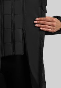 Fekete szigetelt dzseki steppelt párnázással, két elülső cipzárral és sima textúrával, amely egy nyitott oldalt tartó kezet mutat.