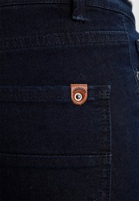 Los jeans de mezclilla oscura presentan una etiqueta de marca de cuero en el bolsillo trasero, con una textura suave y costuras en contraste a lo largo de las costuras.