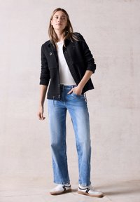 Giacca nera effetto tessuto con chiusura a bottoni, t-shirt bianca, jeans azzurri chiari e sneakers bianche con dettagli neri. Abbigliamento casual.