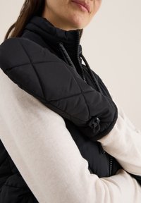 Černá vyp padded vesta s vysokým límcem, s prošívanými detaily na rameni a černými kovovými prvky. Měkká béžová tkanina po straně.