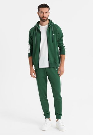Homme debout portant un sweat à capuche zippé vert et un jogging assorti, avec des baskets blanches et un t-shirt blanc, sur un fond uni.