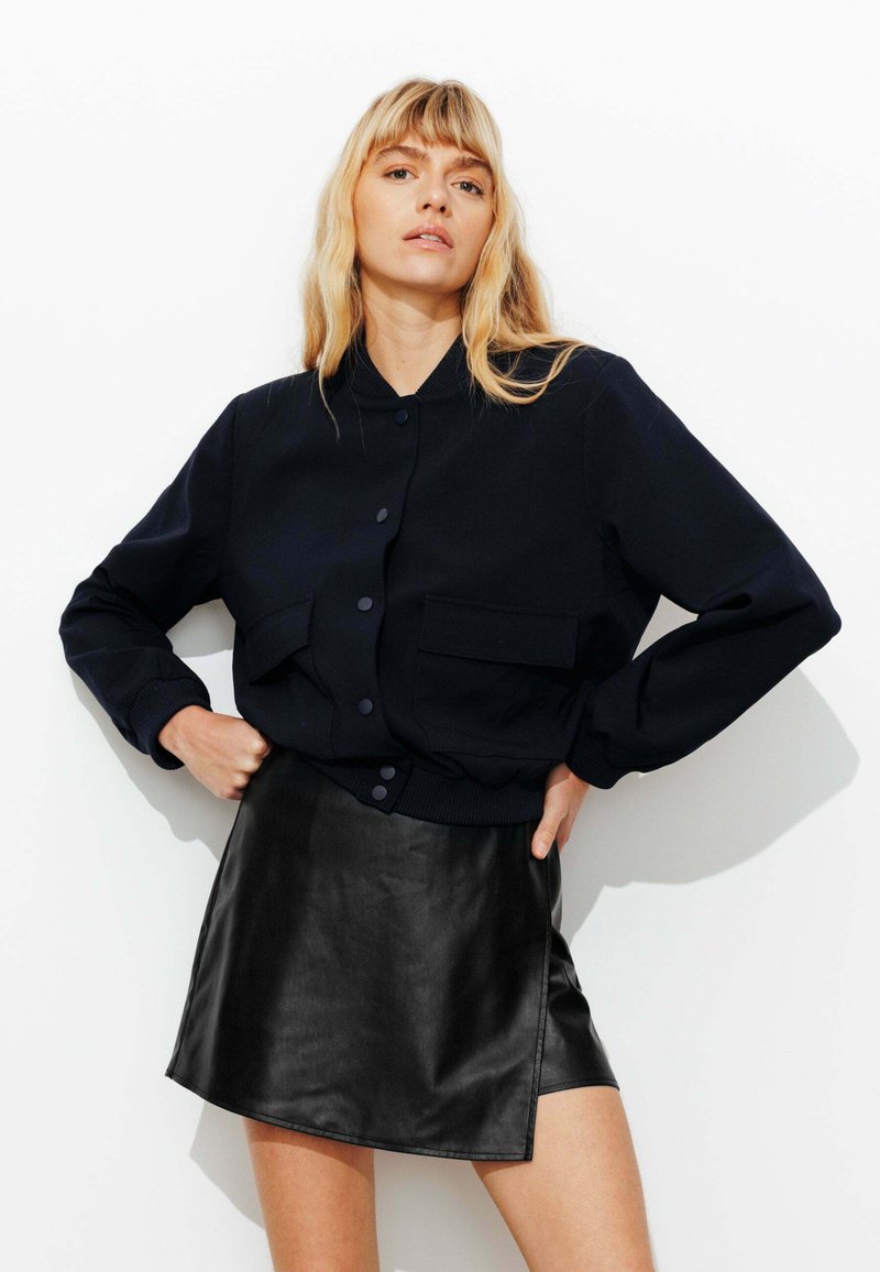 Veste de bombardier navy avec fermeture à boutons, deux poches avant et poignets côtelés. Associée à une mini-jupe en simili cuir noire, texture lisse.