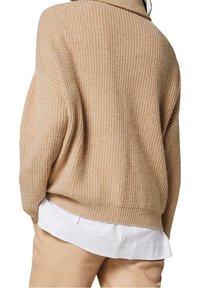 Pull en tricot texturé beige avec une coupe ample, des épaules tombantes et un col roulé, superposé sur une chemise blanche, associé à un pantalon tan.