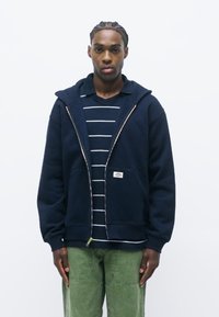 Felpa navy con zip e cappuccio, realizzata in tessuto morbido, con tasca frontale e etichetta con logo. Indossata sopra una maglietta a righe blu navy e bianche.