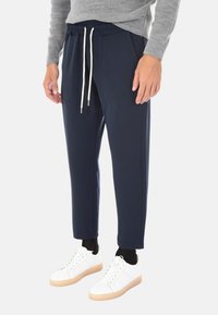 Uomo che indossa pantaloni blu navy con cordoncino, camicia grigia a maniche lunghe, calzini neri e scarpe da ginnastica bianche con suole in gomma, in piedi su uno sfondo bianco.