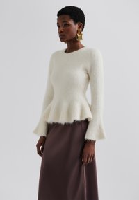 Un pull blanc, moelleux et à manches longues avec un ourlet en peplum, associé à une jupe lisse en marron foncé, mettant en valeur des textures douces et des couleurs contrastées.