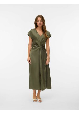 Vrouw staat in een olijfgroene midi-jurk met korte mouwen, geplooide taille en split, gecombineerd met witte open teenhakken, effen witte achtergrond.