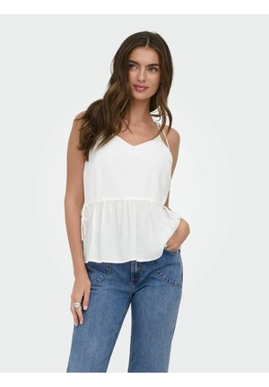 Hosszú barna hajú nő fehér ujjatlan V-nyakú blúzban és kék farmerben, zsebében tartott kézzel pózol.