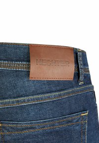 Jeans en denim avec une étiquette en cuir marron affichant "HECHTER." Présente des coutures jaunes et un design de poche arrière. Bords coupés.