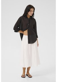 Chemise noire légère à boutons avec des manches retroussées, assortie d'une jupe blanche fluide jusqu'aux genoux et de tongs noires.