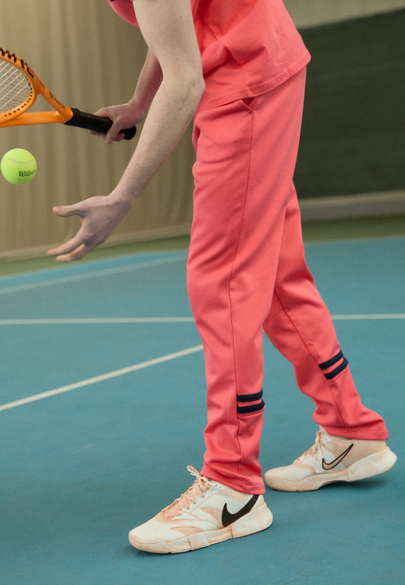 Lysende pink træningsbukser med marineblå striber ved anklerne. Hvide sneakers med et sort Nike-swoosh. En tennisracket holdes, med en gul bold i bevægelse.