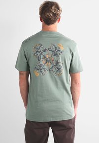 T-shirt en coton vert avec un grand motif floral en bleu, or et noir au dos. Manches courtes et coupe régulière.