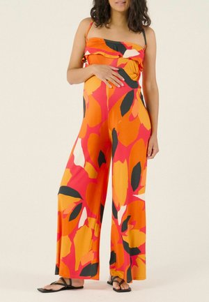 Femme enceinte portant une combinaison sans manches à jambes larges avec des motifs floraux orange, rouge, noir et blanc, chaussée de sandales noires.