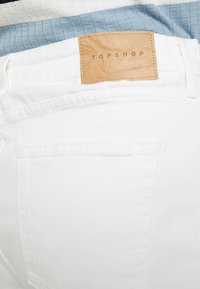 Vita denimshorts med en läderlapp märkt "TOPSHOP" vid midjan, med en slät yta och minimala sömnadsdetaljer.