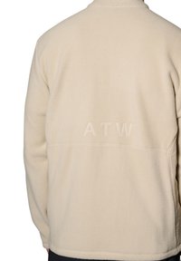 Beige fleecejacka med hög krage, sömnadsdetaljer och en präglad "ATW"-logotyp på baksidan. Mjuk textur och avslappnad passform.