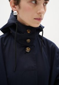 Lietusmētelis tumši zilā krāsā ar augstu apkakli, trim brūnām pogām un aukliņu. Izskatu papildina sudraba sfēriskas auskari.