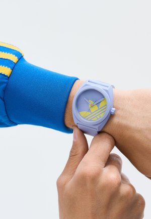 Violets silikona pulkstenis ar apaļu seju, ar dzelteniem akcentiem un svītrām. Nēsāts uz rokas, komplektēts ar zilu svītrainu piedurkni.