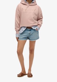 Sudadera rosa con el logo de "SuperDry", llevada sobre una camisa de rayas azules, combinada con pantalones cortos de mezclilla y sandalias marrones con tiras trenzadas.