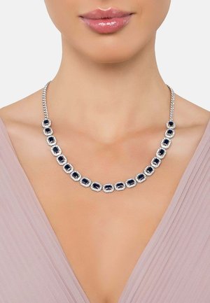 Silver-toned necklace featuring oval blue stones encircled by clear accents, resting on a smooth neckline, against a light fabric background.