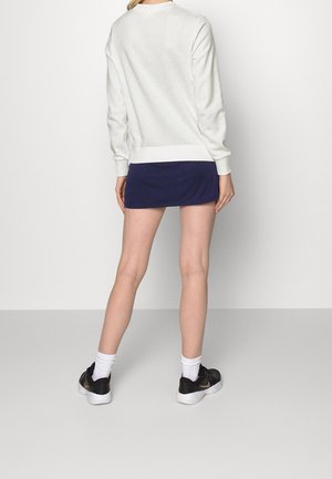 Femme debout, de dos, portant un haut à manches longues blanc, une mini-jupe marine, des chaussettes blanches et des baskets noires à semelles blanches.