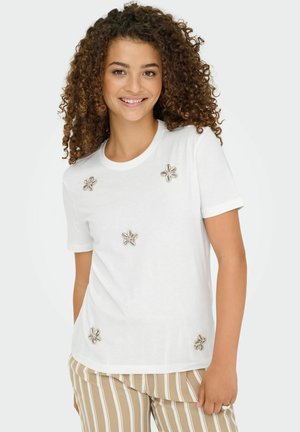 Jeune femme aux cheveux bouclés portant un T-shirt blanc décoré de coques en forme de fleurs et un pantalon rayé beige, souriante.