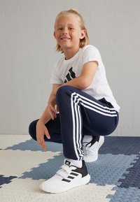 Camisa blanca de manga corta con logo negro, pantalones deportivos azul marino con rayas blancas y zapatillas deportivas en blanco y negro; suelo texturizado multicolor.