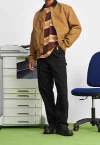 Chaqueta marrón con cremallera y cuello negro, camiseta a rayas burdeos y amarilla, pantalones negros y zapatos negros. Fondo: impresora y silla azul.