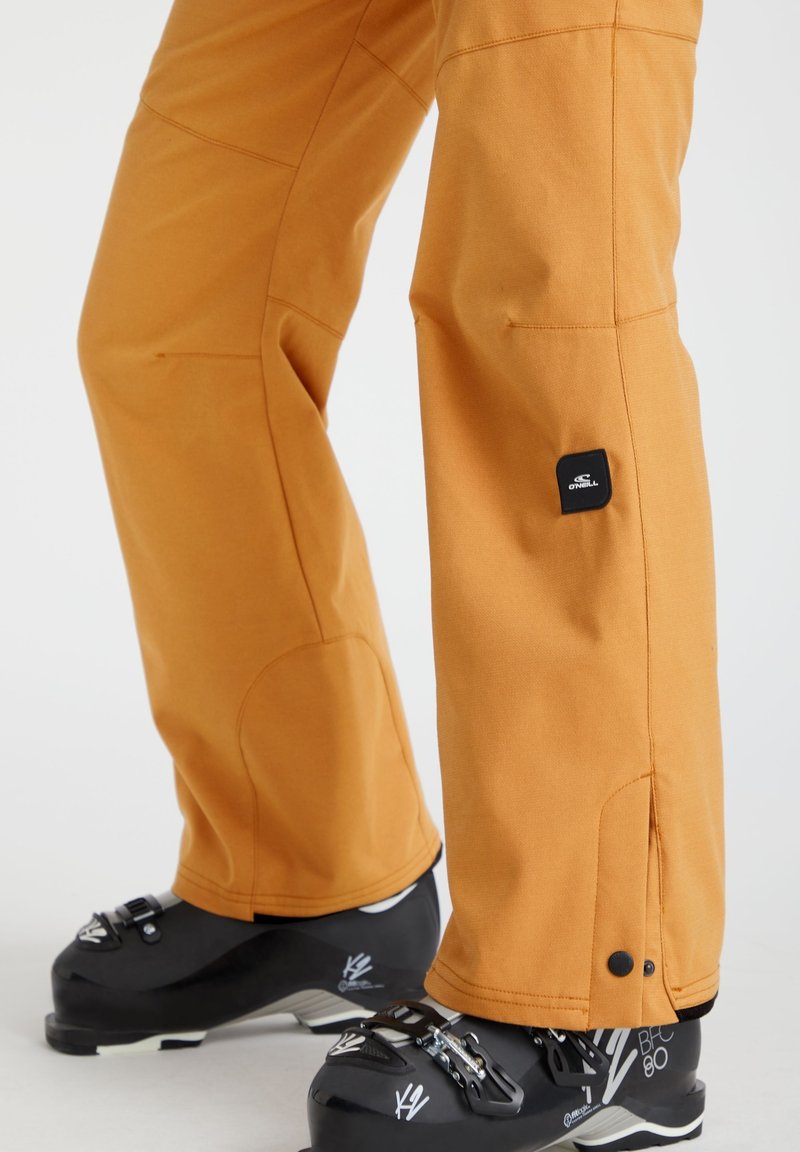 Pomarańczowe spodnie narciarskie z wzmocnieniami na kolanach noszone na czarnych butach narciarskich z klamrami na białym tle.