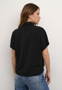 Schwarzes kurzärmeliges Oberteil mit hohem Kragen und glatter Textur, mit einem lässigen Schnitt und einem leichten Glanz. Kombiniert mit blauen Jeans.