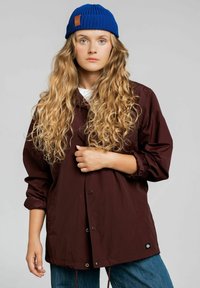 Bordowa lekka kurtka z napami, regulowanym sznurkiem na dole i małą naszywką z logo. W połączeniu z niebieską dzianinową czapką.