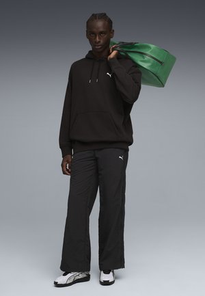 Zwarte hoodie met kangoeroezak en wit logo, gecombineerd met zwarte joggingbroek. Groene sporttas met dubbele handvatten. Zwart-witte sneakers.