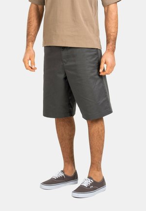 Man wearing charcoal gray knee-length shorts, a beige short-sleeve shirt, and gray lace-up sneakers with white soles.