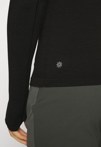 Black long-sleeve shirt with textured fabric and a small, silver logo detail at the hem; worn over gray pants with pockets.