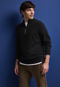 Zwarte sweater met ritssluiting en een zachte gebreide stof, voorzien van een klein embleem op de borst, gecombineerd met bruine corduroy broek tegen een blauwe achtergrond.