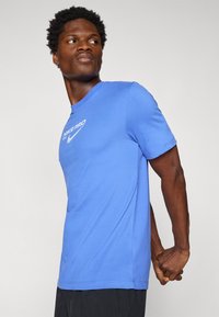 Blå Nike Pro t-skjorte med korte ermer, laget av glatt stoff, med en hvit logo på brystet. Sitter tett inntil kroppen.