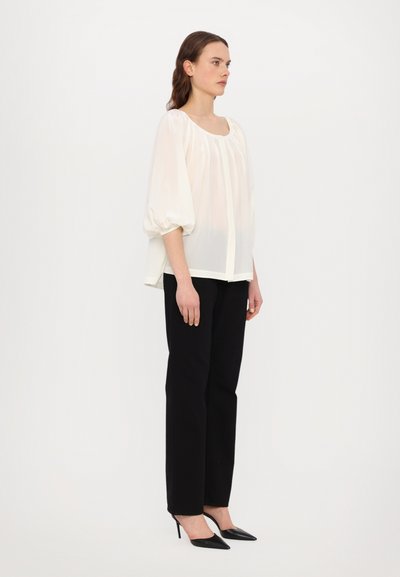 Blouse crème avec un col rond, des manches bouffantes et des plis ; associée à un pantalon noir droit et des escarpins noirs pointus.