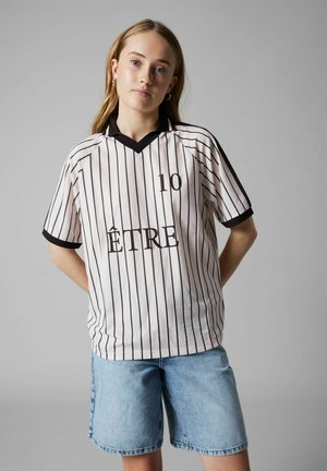 Joven mujer de pie con las manos detrás de la espalda, vistiendo una camiseta blanca con rayas negras, número 10 y la palabra "ÊTRE", y shorts de mezclilla azul claro.