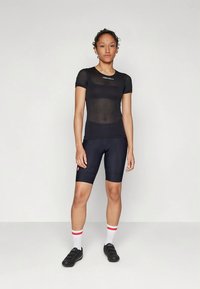 Γυναίκα που στέκεται με μαύρη διχτυωτή μπλούζα ποδηλασίας, μαύρες βερμούδες ποδηλασίας, λευκές κάλτσες με κόκκινες ρίγες και μαύρα παπούτσια ποδηλασίας σε απλό φόντο.