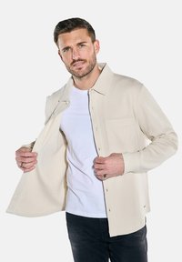 Beige gewobene Hemdjacke mit Knopfverschluss, langen Ärmeln, einer Brusttasche und einem lässigen Schnitt, über einem weißen T-Shirt getragen.