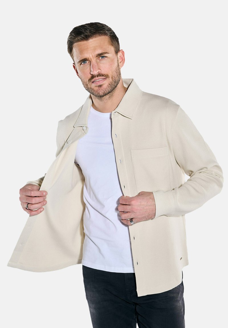 Beige gewobene Hemdjacke mit Knopfverschluss, langen Ärmeln, einer Brusttasche und einem lässigen Schnitt, über einem weißen T-Shirt getragen.