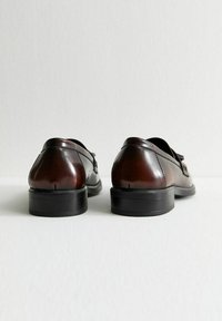 Brązowe skórzane buty z wypolerowanym wykończeniem, zaokrąglone noski i niska czarna gumowa podeszwa. Charakteryzują się prostym wzornictwem z minimalnymi detalami.