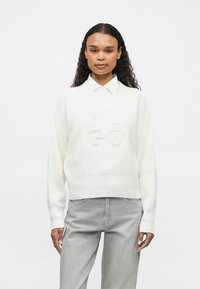 Γυναίκα με μακριά σγουρά μαλλιά, ντυμένη με ένα λευκό πουλόβερ HUGO πάνω από ένα λευκό πουκάμισο και ανοιχτό γκρι τζιν, στέκεται μπροστά σε ένα απλό φόντο.