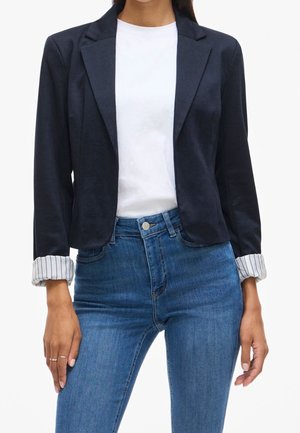 Femme portant un blazer bleu marine foncé avec doublure rayée aux poignets, t-shirt blanc et jean bleu taille haute, debout les bras détendus le long du corps.