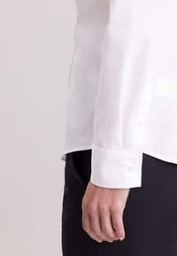 Biała koszula na guziki z gładkiego, lekkiego materiału. Posiada zaostrzony kołnierzyk, długie rękawy oraz mankiety zapinane na guziki.