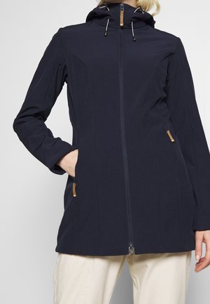 Personne portant une veste à capuche bleu marine avec une fermeture éclair à l'avant et des poches latérales, assortie à un pantalon beige clair, sur un fond uni.